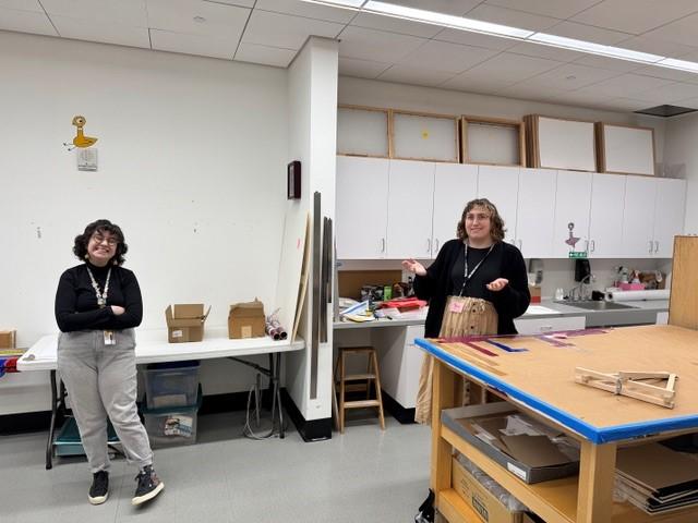 two people in a room with cupboards and boxes in the background and a counter height table with a small easel and framing supplies