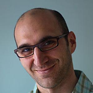 A man wearing glasses in a blue shirt in front of a blue background. 