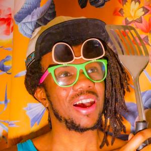 a Black man wearing a hat and sunglasses, and holding a spatula