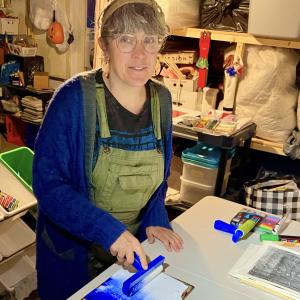 Picture of the artist Angela Zammarelli who is adding blue and white ink to a printmaking plate and is working in a studio space filled with a variety of art materials on shelves. 