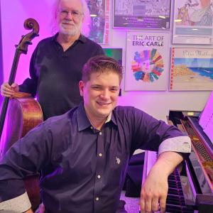 One person sits at a piano and the other stands behind them with a bass.