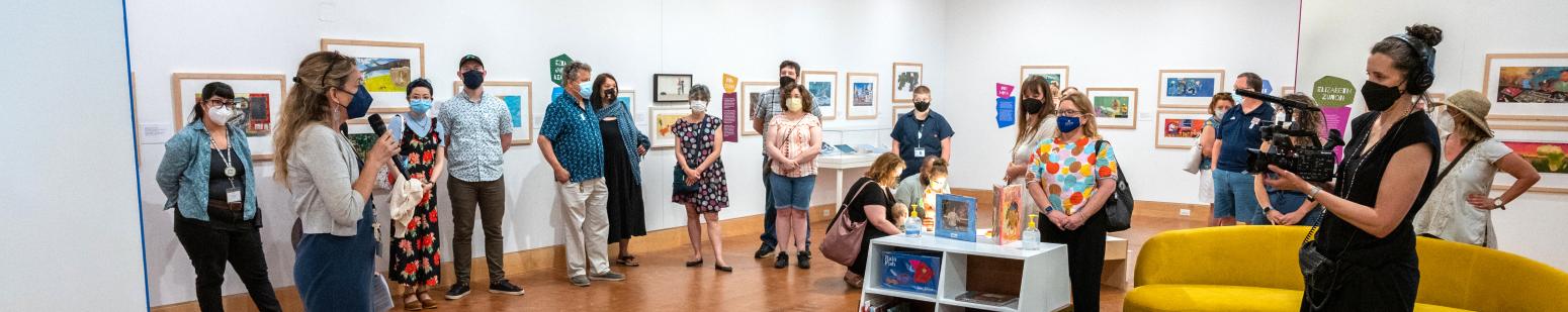 Filmaker filming a group of people standing in art gallery listening to speaker