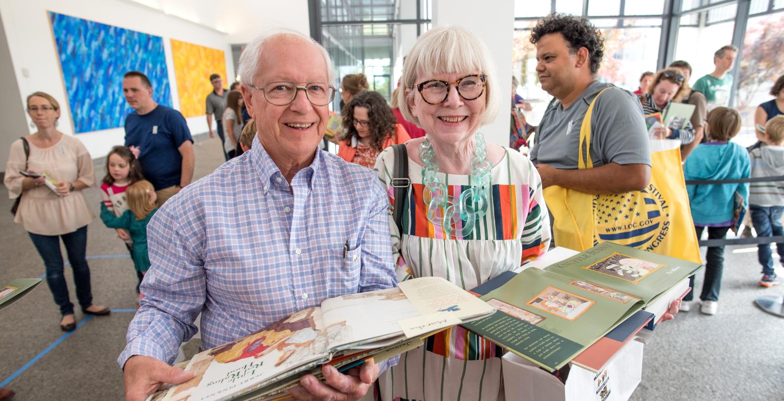 Two members stand in the Great Hall carrying open picture books that have just been signed by an artist or author at an event.