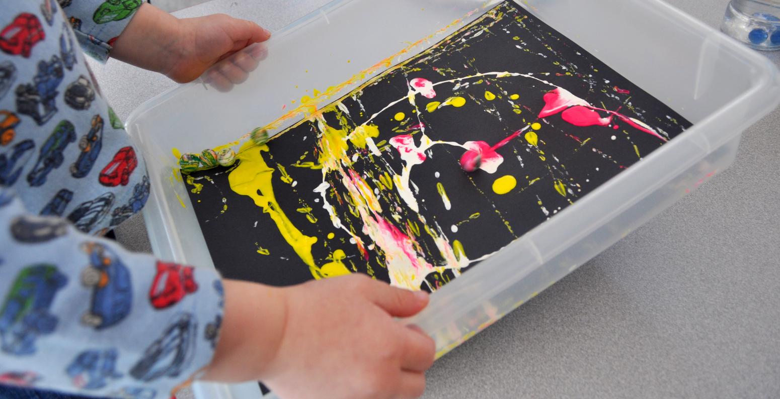 A toddler moving a plastic bin around with a painty marble and black paper inside, creating a colorful print.
