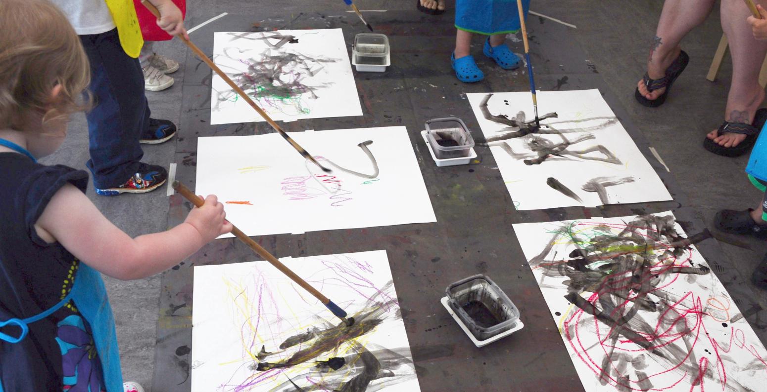 Young children standing and painting with black paint on the floor using long brushes.