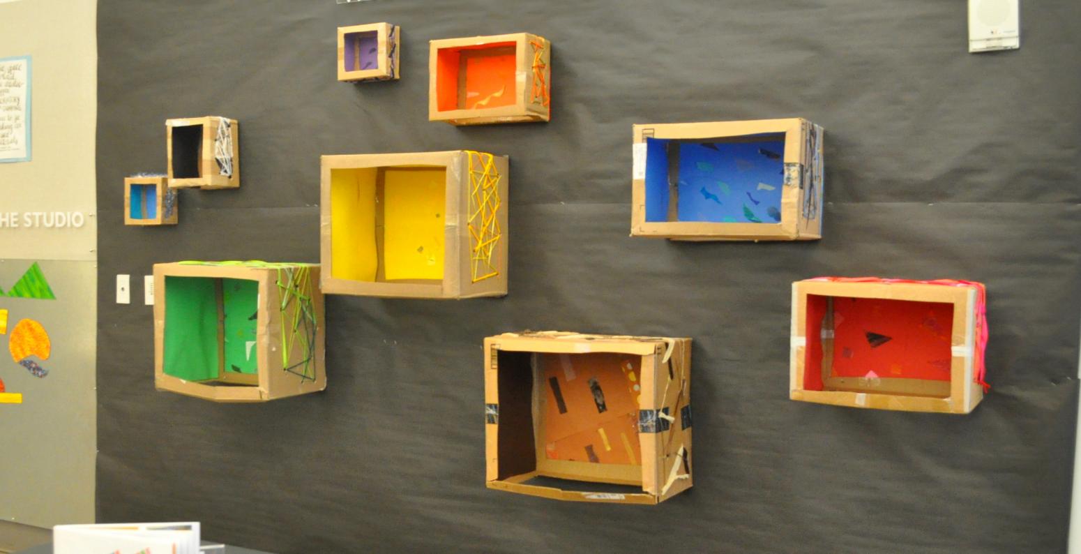 Eight cardboard puppet theaters attached to a display wall, each with a different colored background inside.