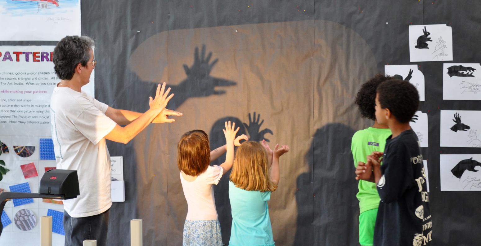 An adult and several children making shadow hand puppets on the wall.