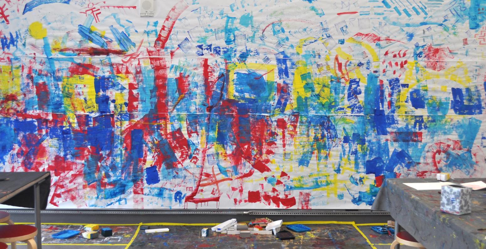 A mural with red, yellow, and blue paint, created by printing with boxes on a white paper backdrop.