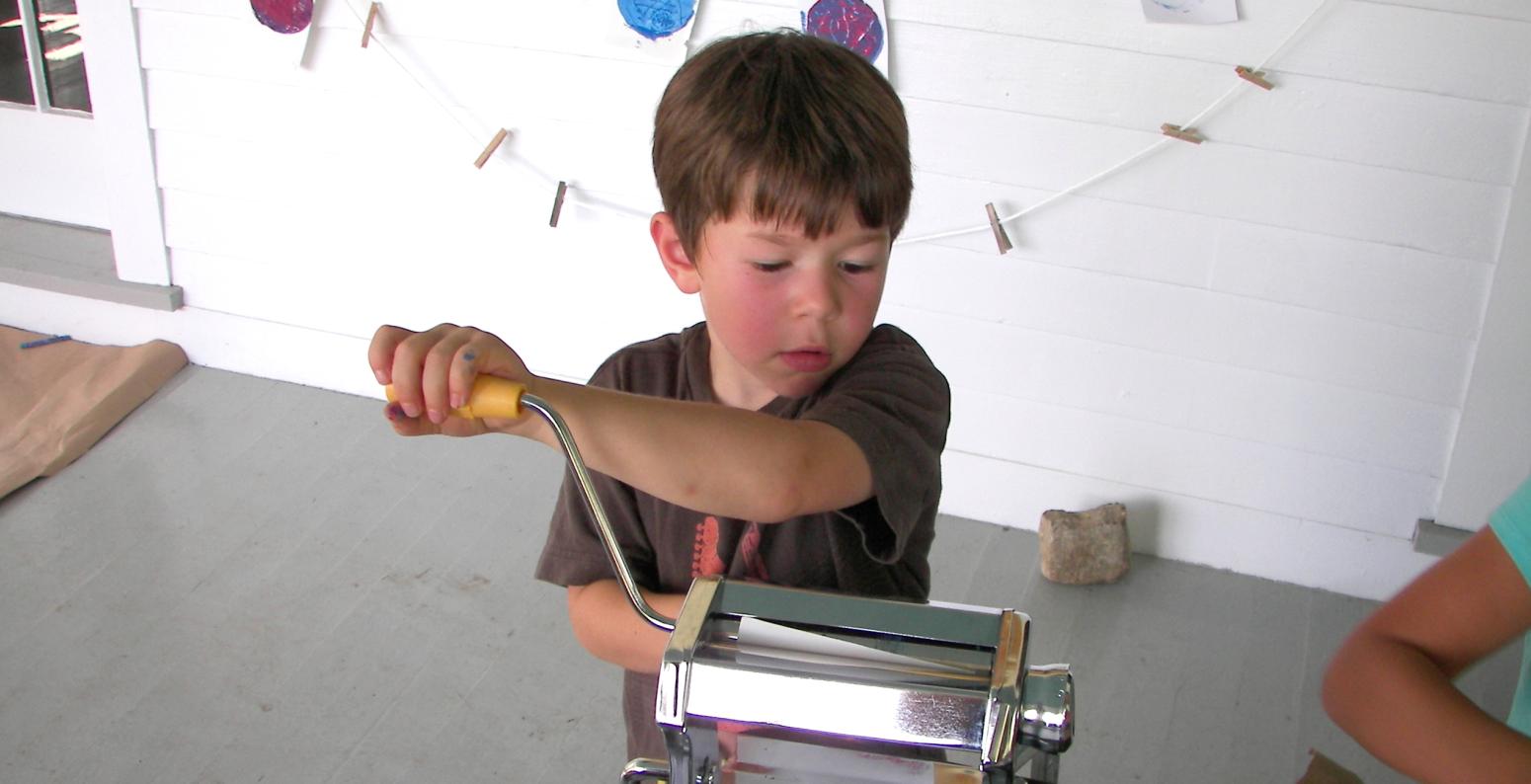 A young boy cranks a pasta machine, using it as a printmaking tool to make a print.