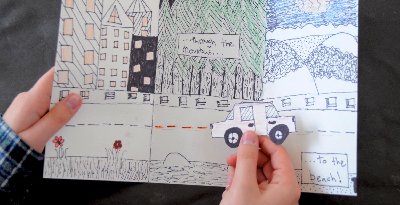 A hand holding a drawing with a road and various landscapes, with another hand holding a paper car in front as if it were driving along the road.