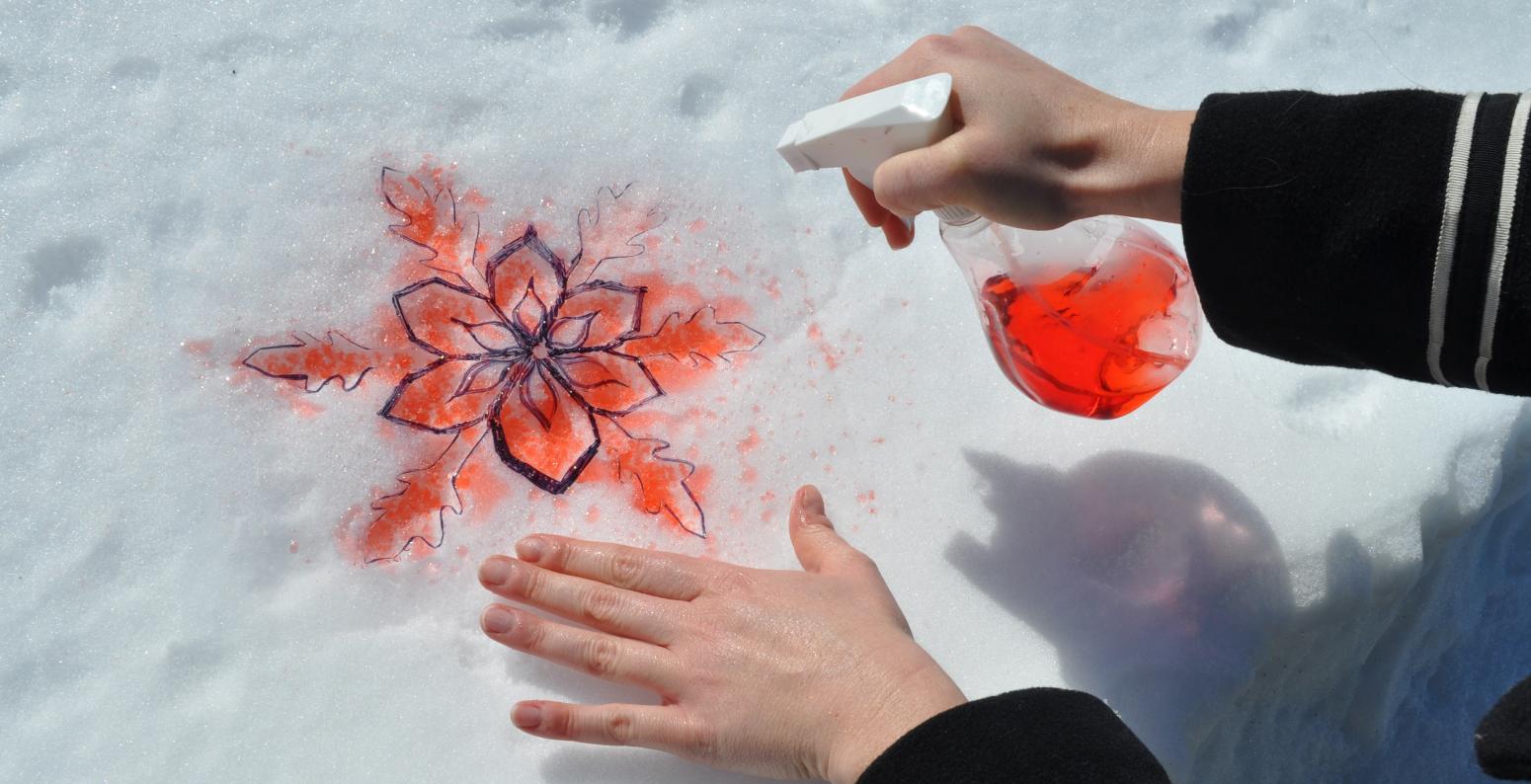 An adult sprays red food-colored-dyed water onto a stencil held onto a pile of white snow.