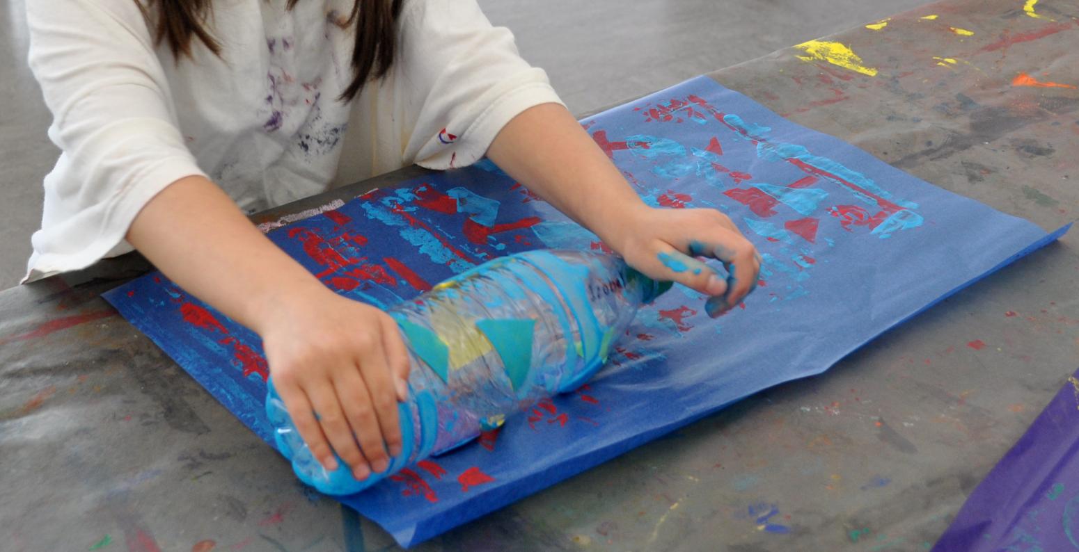 A child rolls a paint-water bottle across a blue piece of paper, creating pattern stamps on it.