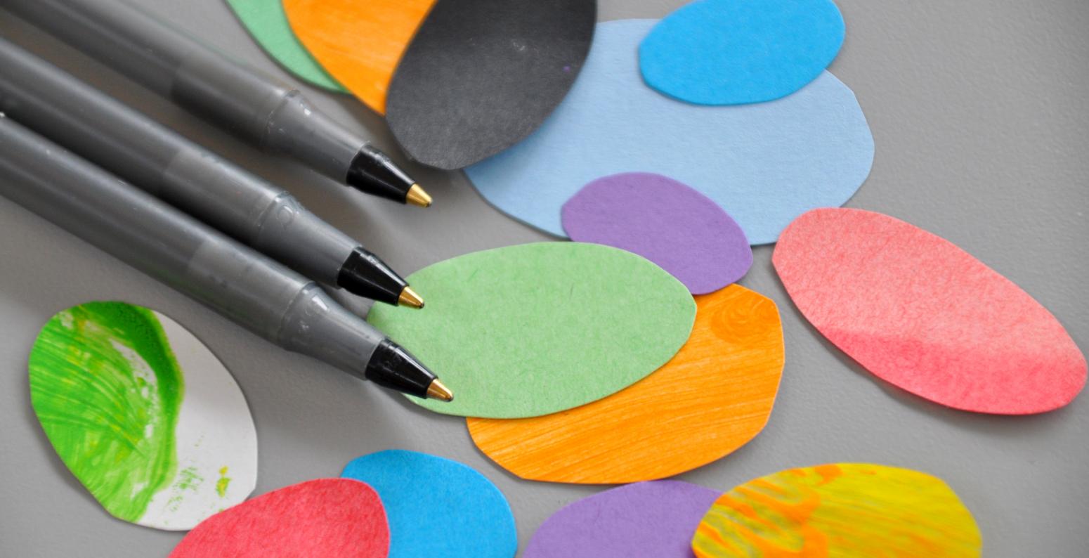 Three black ballpoint pens next to a scattering of brightly colored paper ovals.