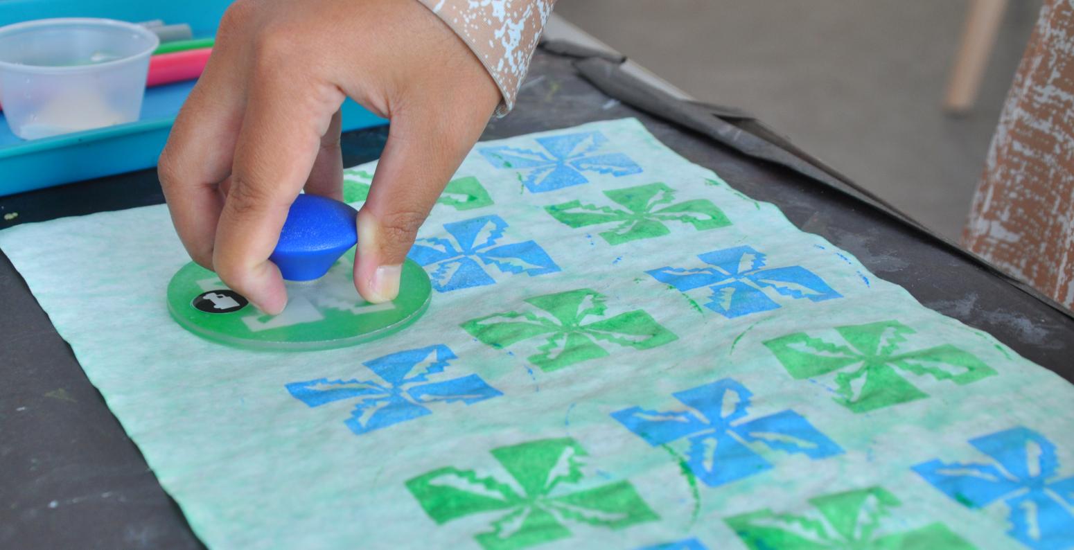 A hand presses a painted stamp onto a piece of dyed paper, creating a pattern inspired by batik.