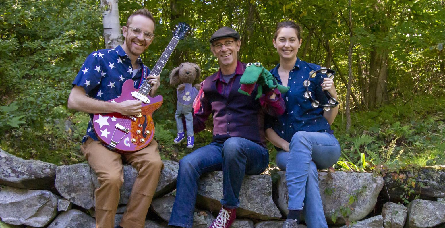 Three musicians sitting on a stone wall with puppets, a guitar, and tamborine.