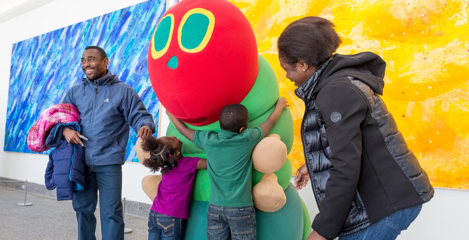 Two children hug The Very Hungry Caterpillar costume character, with two caregivers smiling and standing nearby.