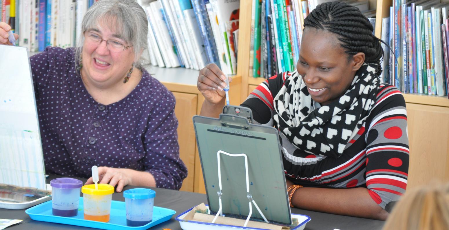 Two seated adults smiling while applying watercolor to upright papers.