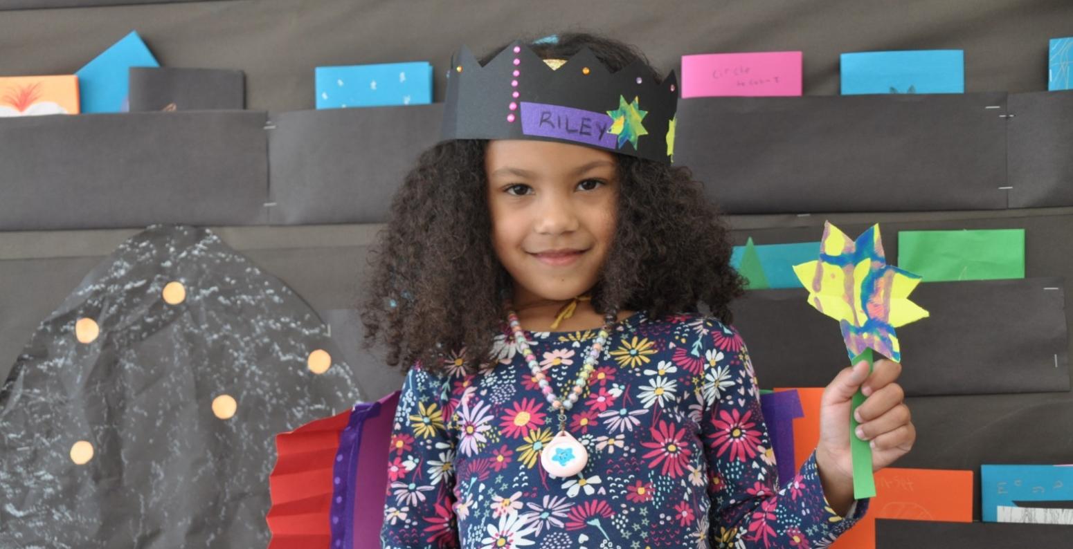 A young artist shows off their paper creations. A bright star shaped wand is held up with pride, a rainbow paper cape rests on the shoulders and a star crown sits on top of the head.