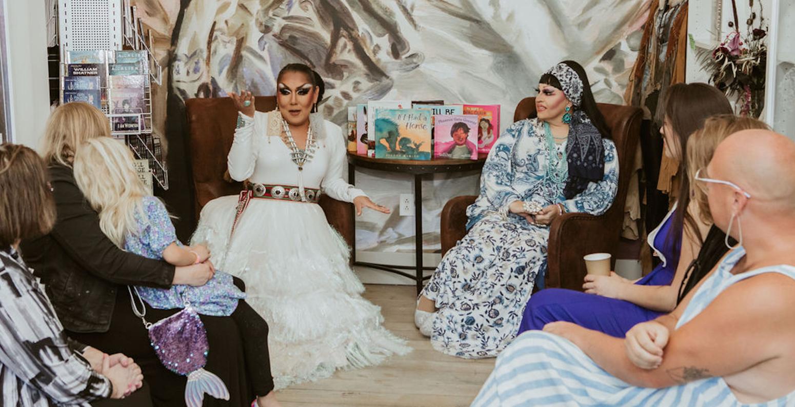 Two drag storytellers sit next to a display of picture books for storytime.