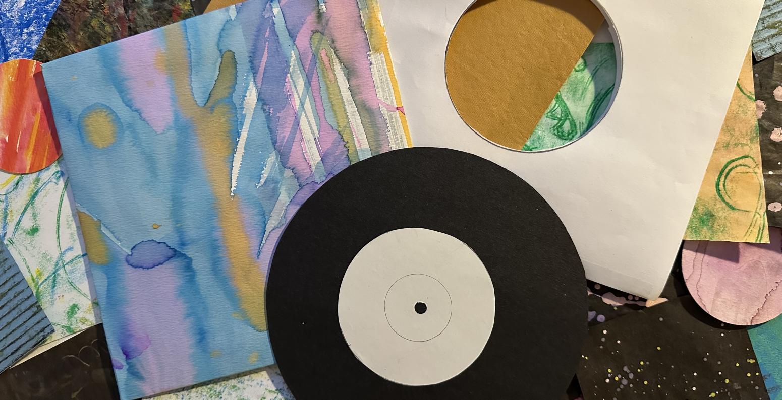 A variety of colorful collage papers in the background with a vinyl record cut out of black card stock laying on top of a blank white record inner sleeve and a colorful record jacket with light blue, pink, and peach paint.