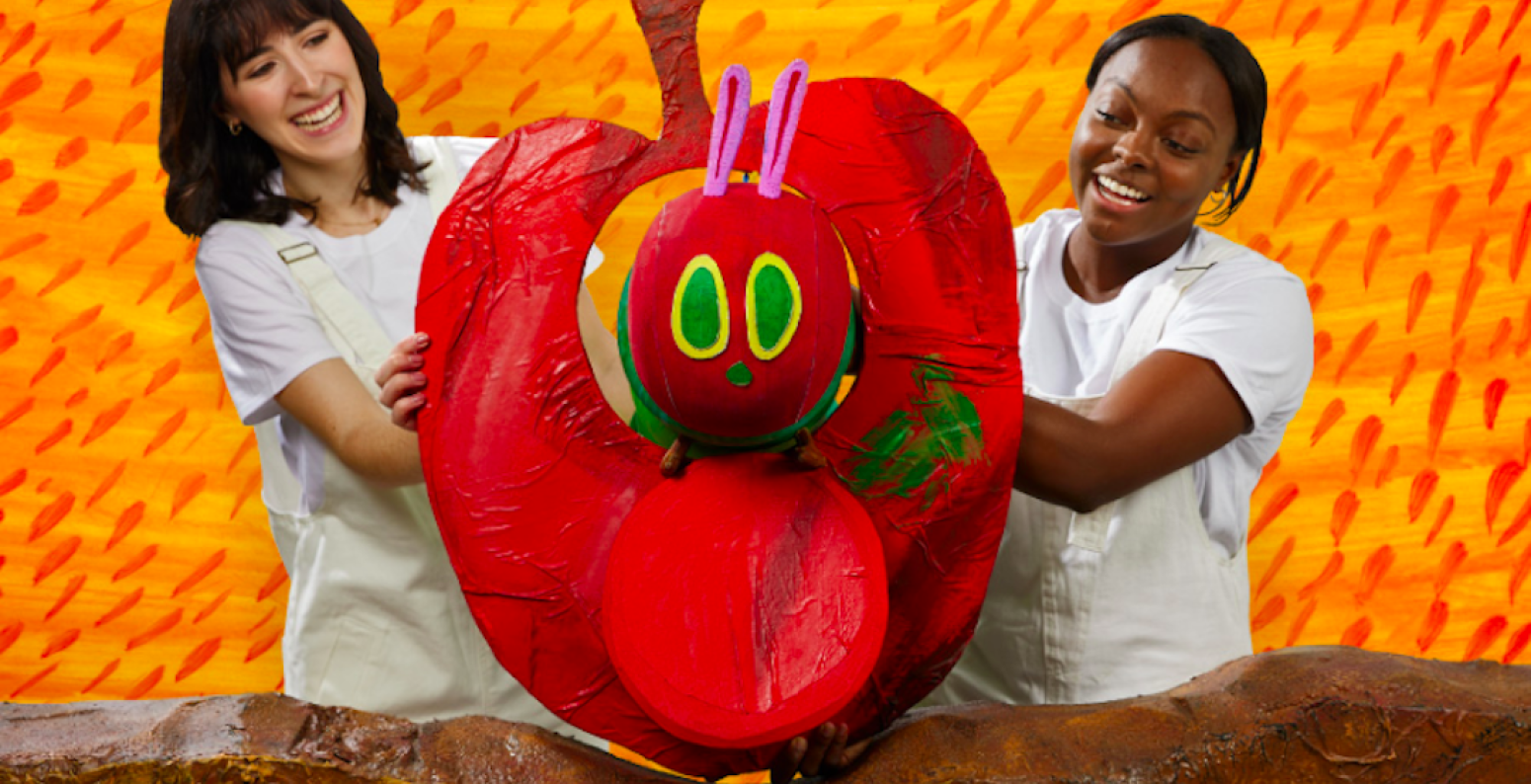 Two puppeteers smiling with a Very Hungry Caterpillar puppet eating through a large red apple.