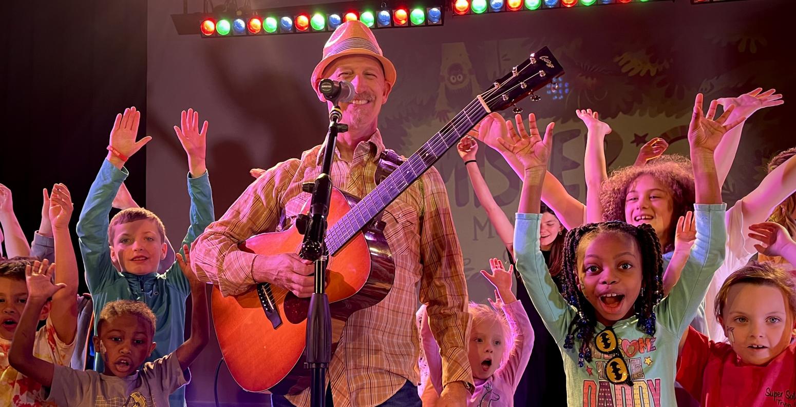 Mister G holding a guitar surrounded by children with their hands raised in the air.