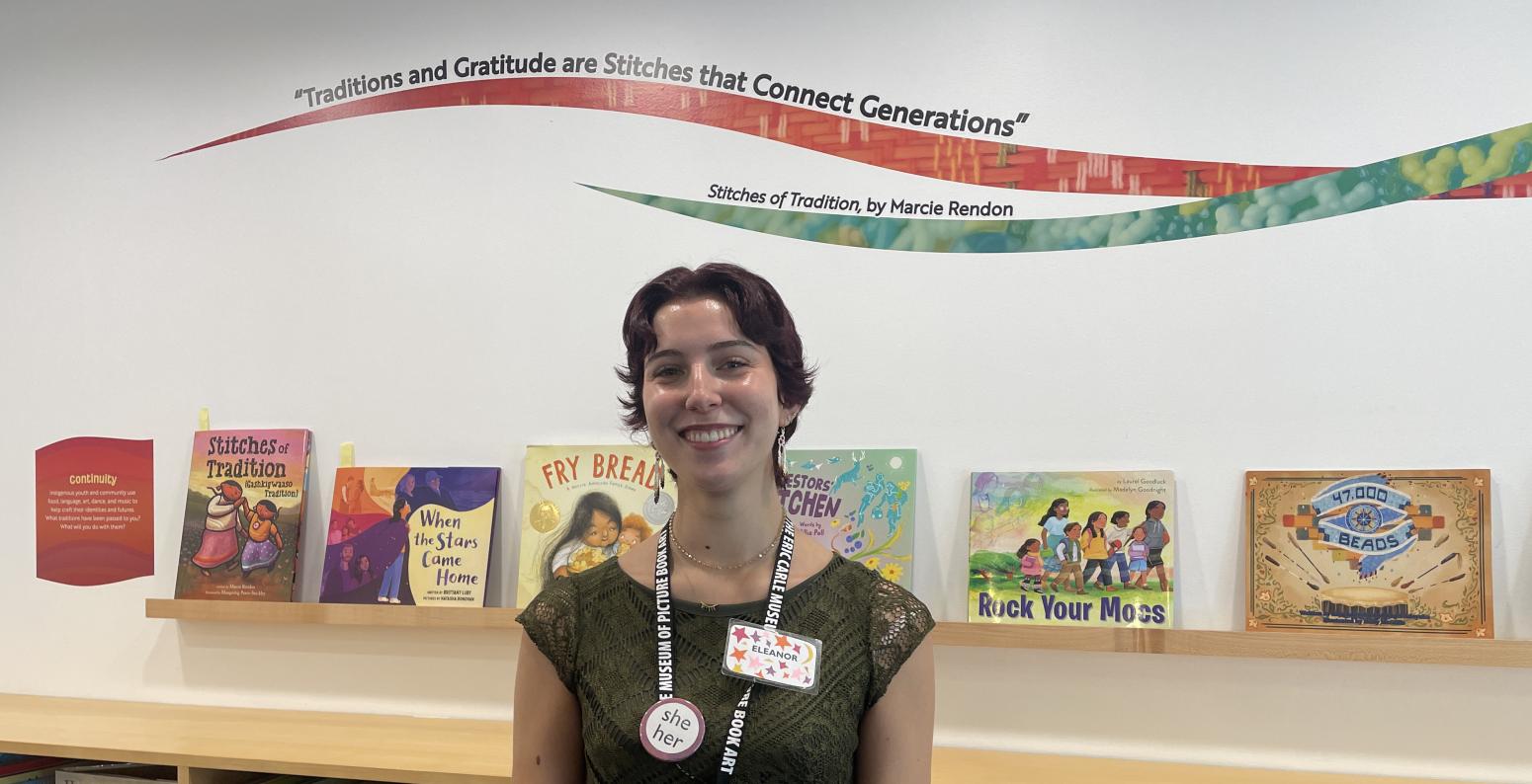 Young woman with short brown hair with a Museum lanyard around her neck and picture book exhibition in the background 