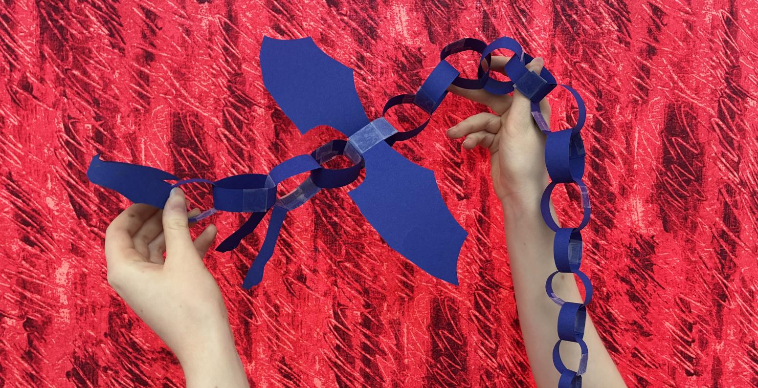 A blue paper chain dragon against a red fabric background. hands holding the paper creation up. 