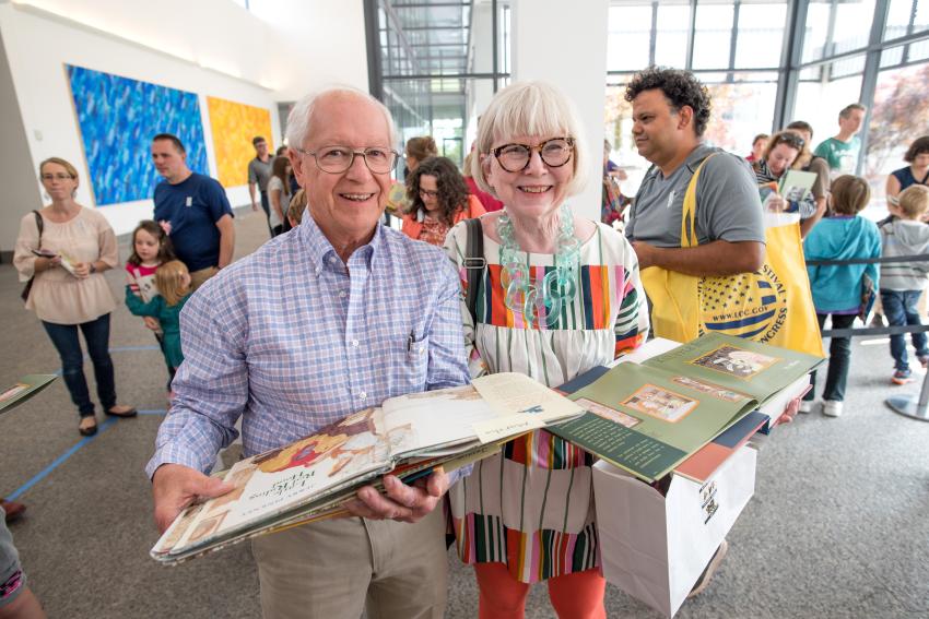 Two members stand in the Great Hall carrying open picture books that have just been signed by an artist or author at an event. 