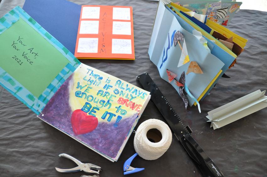 The three books with string, two hole punchers, a crown binding, and a long reach stapler. The Your Art Your Voice book is open to the first page.