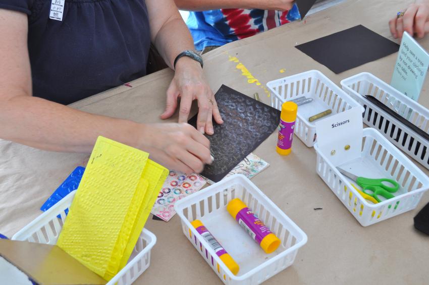 An adult uses a rubbing plate to add texture to their drawing on black paper.