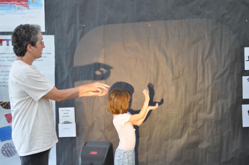 An adult and a child make shadow puppets on a large black bulletin board.