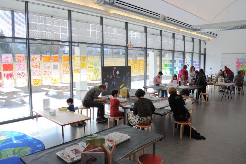 Art studio with guests at 4 tables making art. The tables have supplies and books on them.  A window display with colorful paintings.  