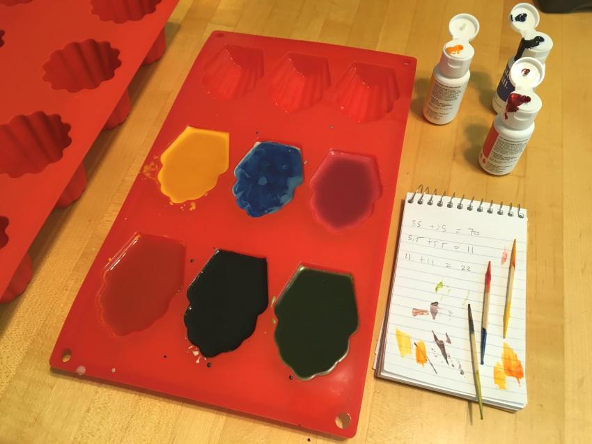 Six colorful crayons cooling in a red mold next to the food colorant bottles, a notebook, and toothpicks used for stirring.