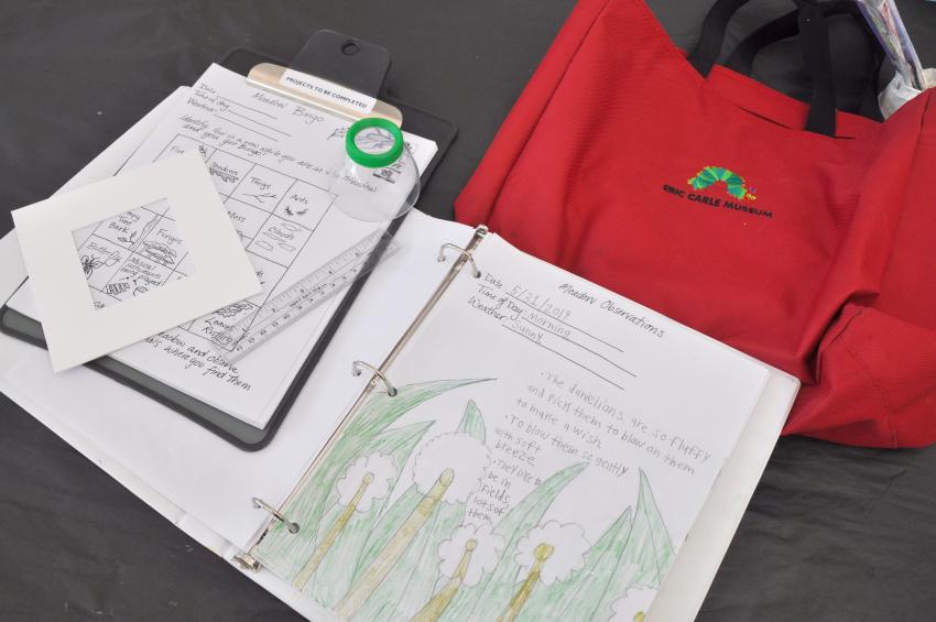 A red bag next to a clipboard with a meadow scavenger hunt, a magnifying lens, and a meadow observation drawing.