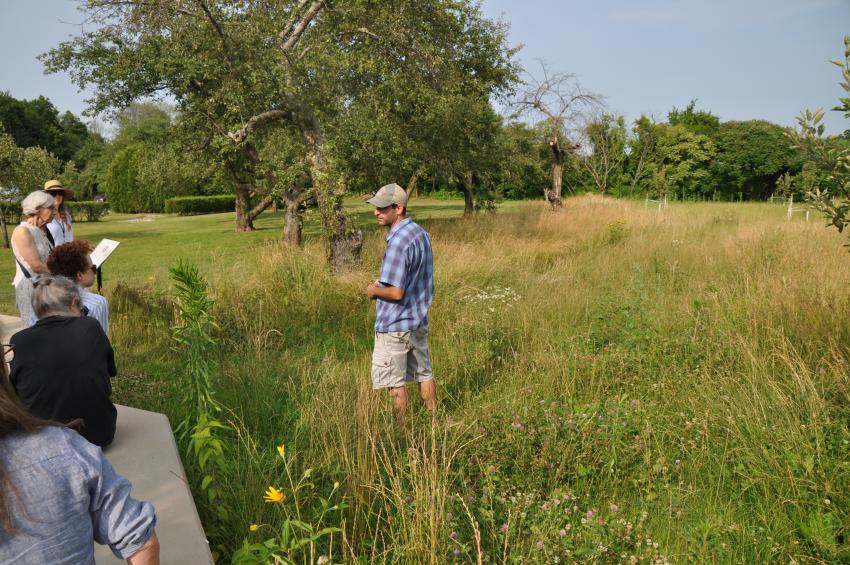 Meadow consultant Owen Wormser stands in the meadow and speaks with volunteers sitting and standing on the paved meadow path.