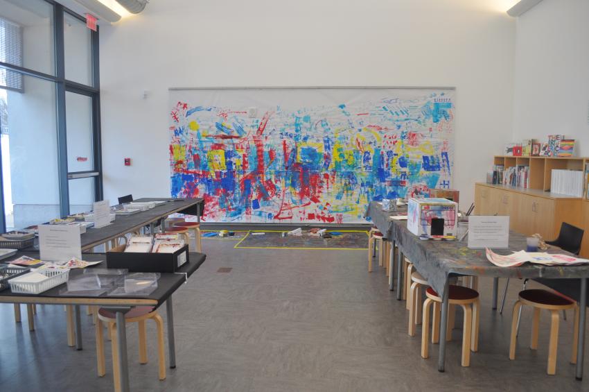 An empty Art Studio with materials in baskets on the tables and a large, painted, collaborative mural in the background.