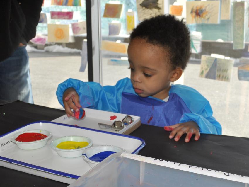 A young child wearing a smock gently places a small that was rolled into paint into a shallow box.