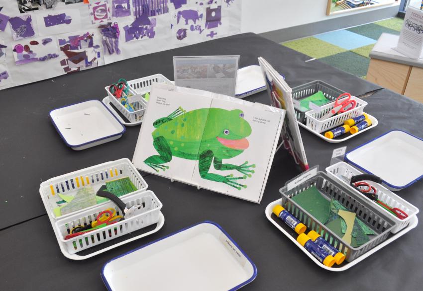 An Art Studio table with art materials, scrap trays, and the Brown Bear book open to the green frog page. 