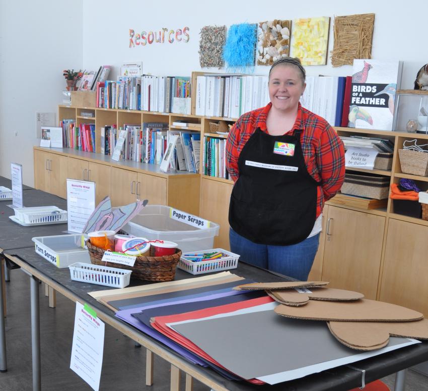 Intern Rebekah smiles and stands in front of a table of art materials in the Art Studio.