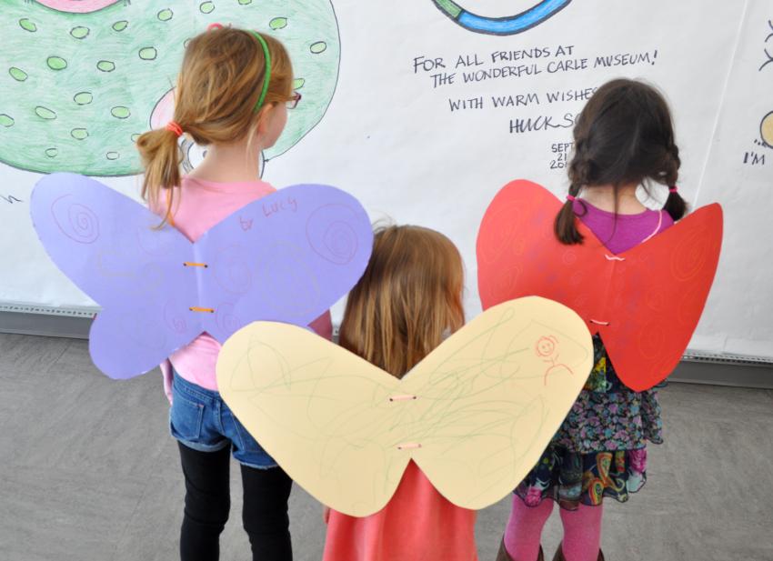 Three children wearing their own handmade paper wings, each in a different color and with different designs on them.