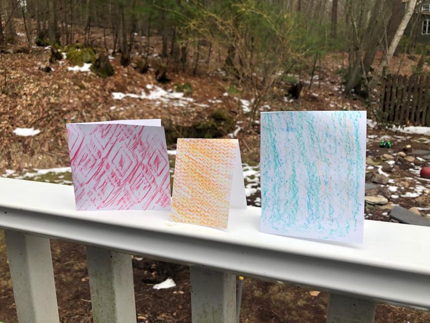 Three cards, with texture patterns on them, sitting on a porch rail outside.