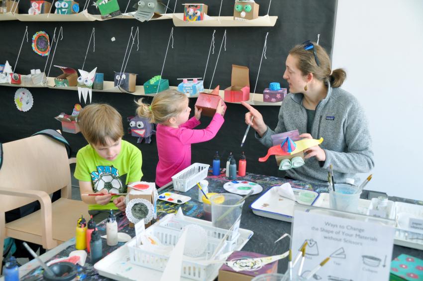 A family works together to make their own sculptures with a sign in the middle of the table suggesting ways to change the shape of their paper without scissors.