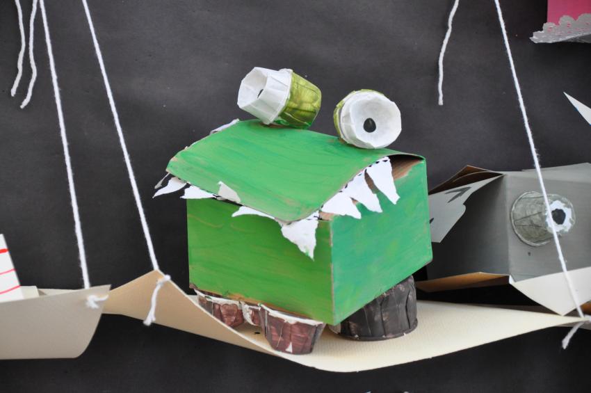 A crocodile sculpture with torn paper teeth, cups for feet, and eyes made from cups.