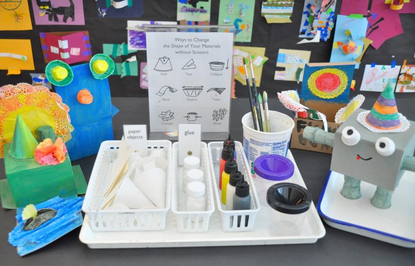 A tray of materials, sample sculptures, and a sign that suggests different ways to change the shape of paper without scissors.