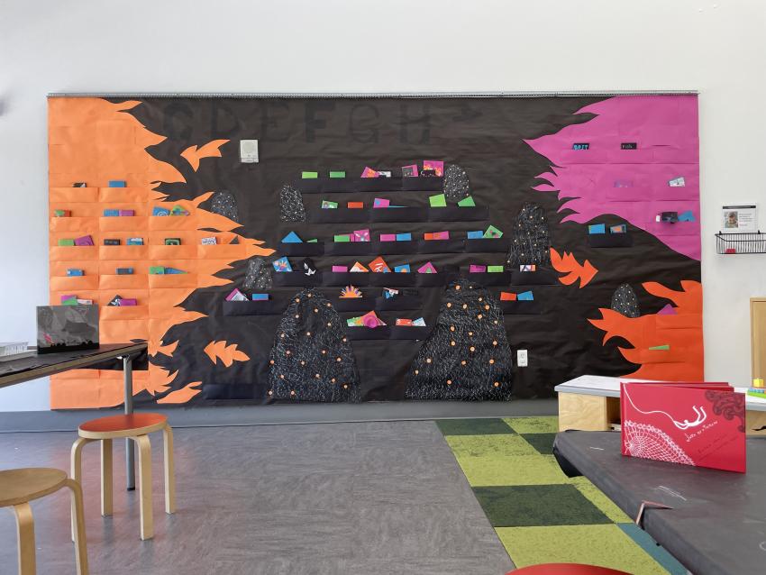 Display wall in the studio. Pink, neon orange, and red orange flamed paper cut outs on the right and left side of the wall. Colorful books pop out of paper pockets and are tucked behind black paper rocks that have bright orange dots on them.