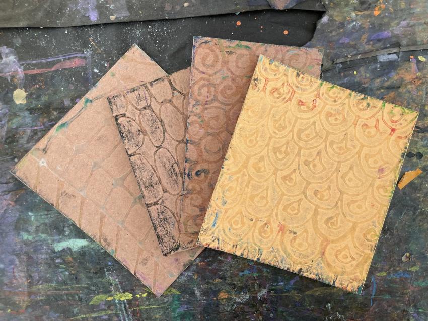 Four sheets of thick paper with designs drawn in glue that have dried to be used as textured rubbing plates.