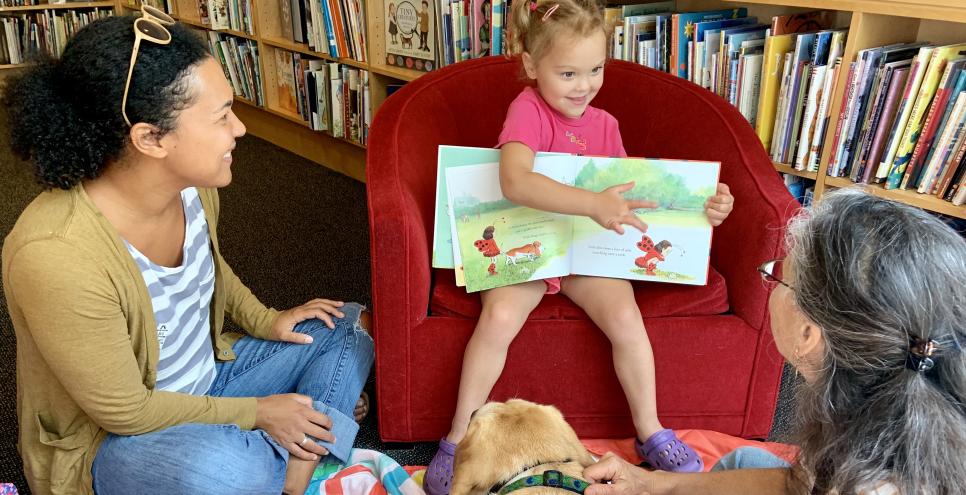 Child sitting in chair showing picture book to reading buddy dog.