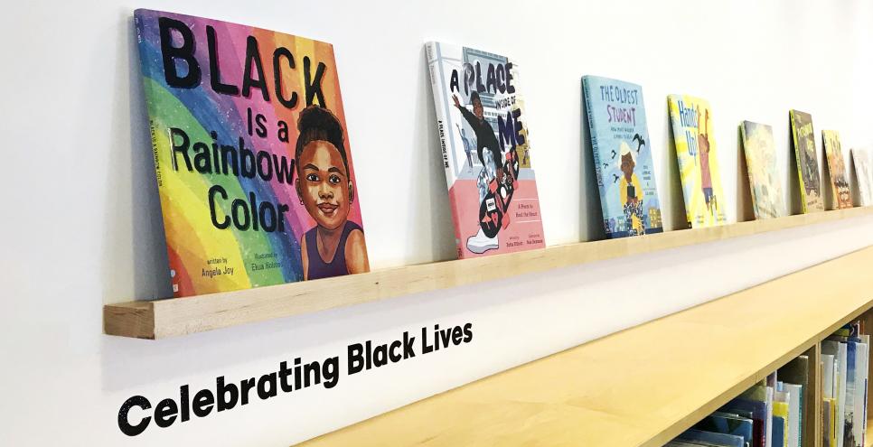 Colorful books sit on a wood ledge against a white wall. The black text on the wall reads "Celebrating Black Lives."