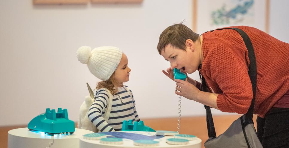 Adult with a surprised expression answers a telephone in an interactive display in the West Gallery as child in hat is amused 